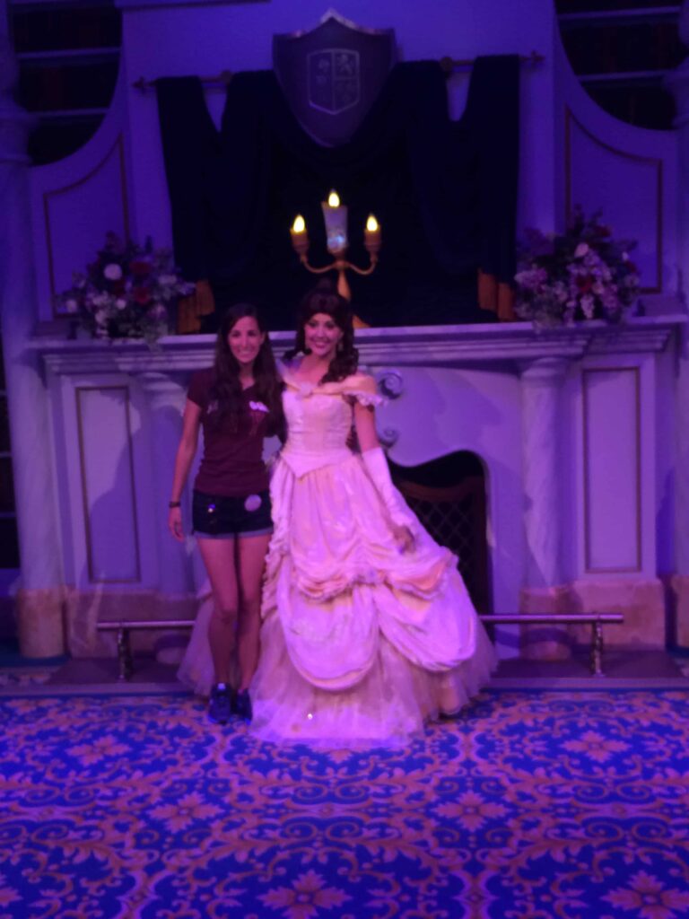 Mulher ao lado da Princesa Bela de A Bela e a Fera com Lumiere, também do mesmo filme, ao fundo, dentro da atração "Enchanted Tales with Belle" o parque de diversões Magic Kingdom no Walt Disney World Resort