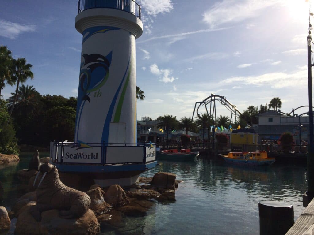 Entrada do parque SeaWorld Orlando, onde é possível ver um farol com uma baleia pintada, esculturas em pedra de leões marinhos e mais ao fundo, barcos e montanha russa