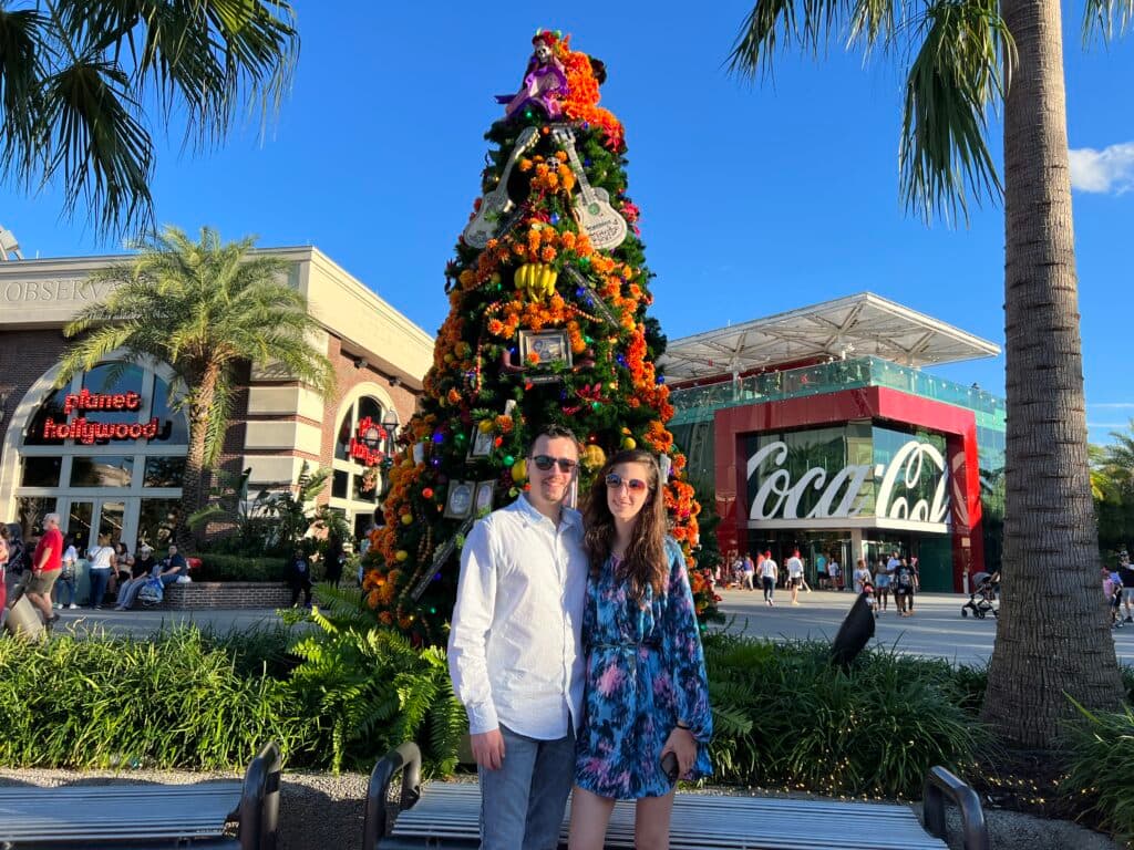 Foto que mostra um casal sorridente, bem vestido e de óculos de sol em frente a uma árvore de natal decorada com o tema do filme VIVA da Disney Pixar. Mais ao fundo é possível ver também o restaurante Planet Hollywood e a loja oficial da Coca Cola, todos localizados em Disney Springs