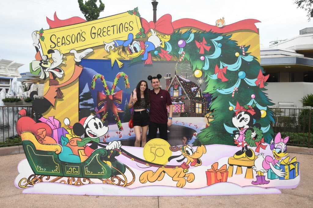 Foto divertida em um pop up decorativo com tema natalino. Casal ao centro e ao seu redor Mickey em um trenó puxado por seu cachorro Pluto, leva sacos cheios de presentes. Minnie e Margarida enfeitam a base da árvore de natal com presentes e laços, enquanto Pato Donald no topo da árvore tenta posicionar a estrela e o Pateta coloca uma placa de Boas Festas, fazendo referência a festa de Natal do Magic Kingdom, na Disney