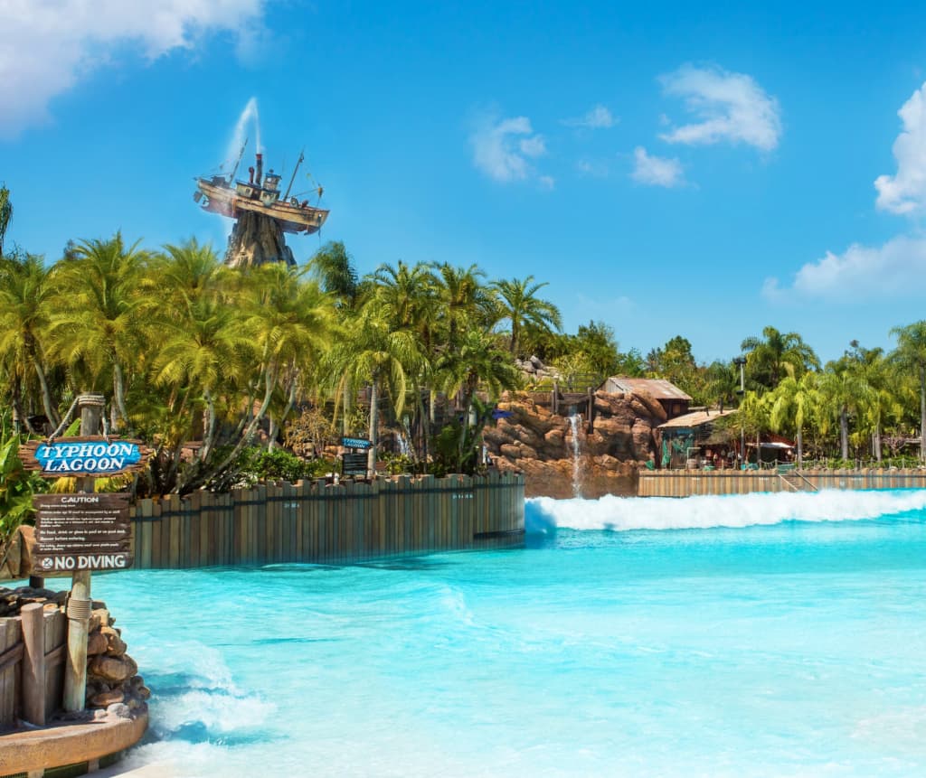 Foto do parque aquático da Disney, Typhoon Lagoon, com seu barco preso na montanha, símbolo da história do parque, que nasceu de um tufão. A frente a água limpa e cristalina da piscina de ondas, com diversas paineiras ao redor, e mais ao fundo, o céu azul de um lindo dia