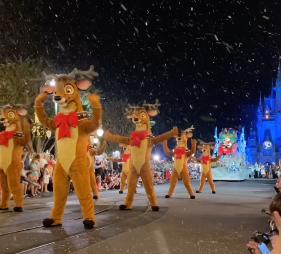 Renas dançam para o público que assiste ao desfile da festa de natal da Disney. Ao fundo é possível ver o Papai Noel chegando em seu trenó e ainda mais ao fundo o Castelo da Cinderela. Claro, também está caindo neve para deixar o clima de natal ainda mais perfeito.