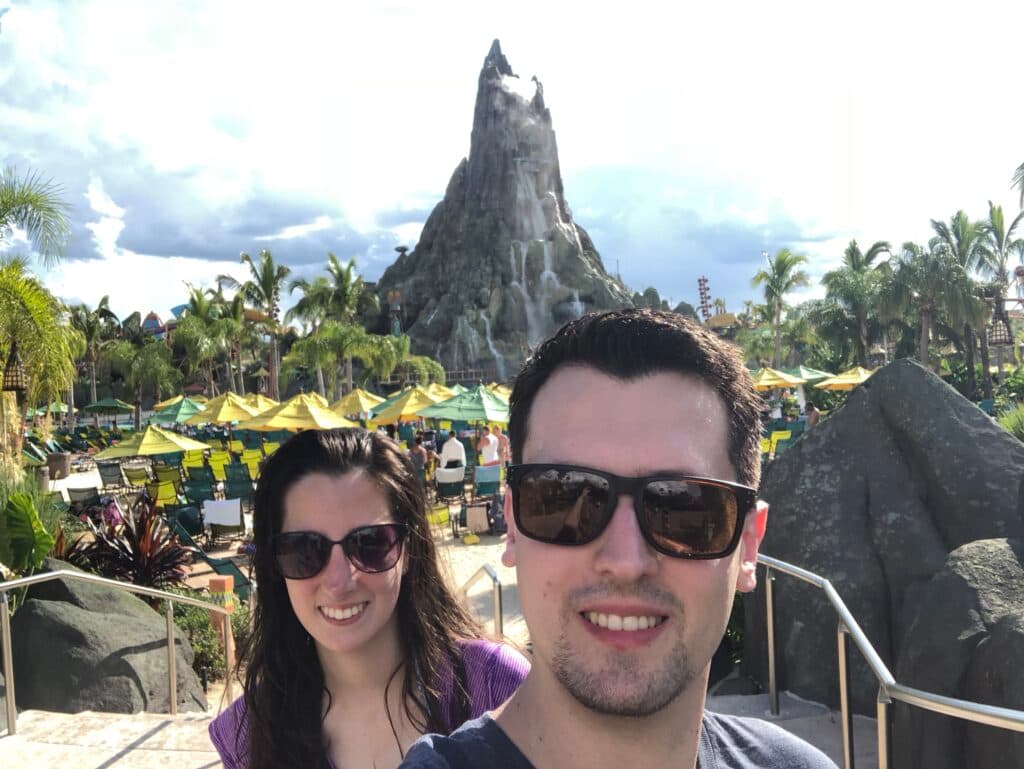 Casal jovem em frente ao Vulcão do parque aquático Volcano Bay, na Universal Studios Orlando. Ambos usam óculos de sol e tem os cabelos molhados. Ao fundo é possível ver diversos guarda sois nas cores verde e amarelo assim como cadeiras espreguiçadeiras