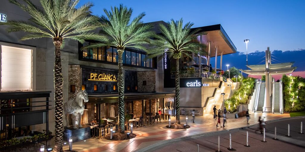 Lojas e restaurantes do shopping Mall At Millenia em Orlando. É possível ver um céu azul escuro de fim de dia, palmeiras grandes e iluminadas, escadas rolantes e a linda arquitetura do centro de compras