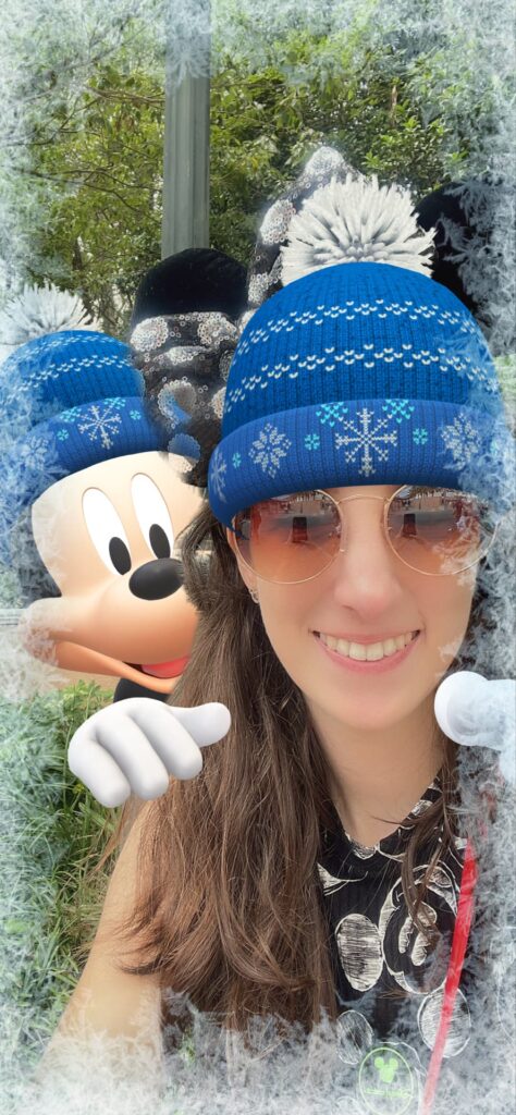 Menina em foto estilo selfie usando um gorro de frio azul com flocos de neve, que está combinando com o gorro do mickey, que aparece em realidade aumentada abraçando-a por trás.