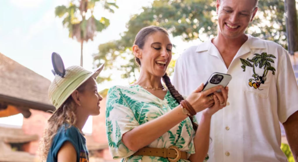 Foto mostra um casal com uma menina, que usa um chapéu com orelhas do Mickey, personagem que também está presente na camisa do homem. A mulher segura um celular para qual todos estão olhando e sorrindo, simbolizando quanto o uso do Genie+ facilita a experiência nos parques Walt Disney World.