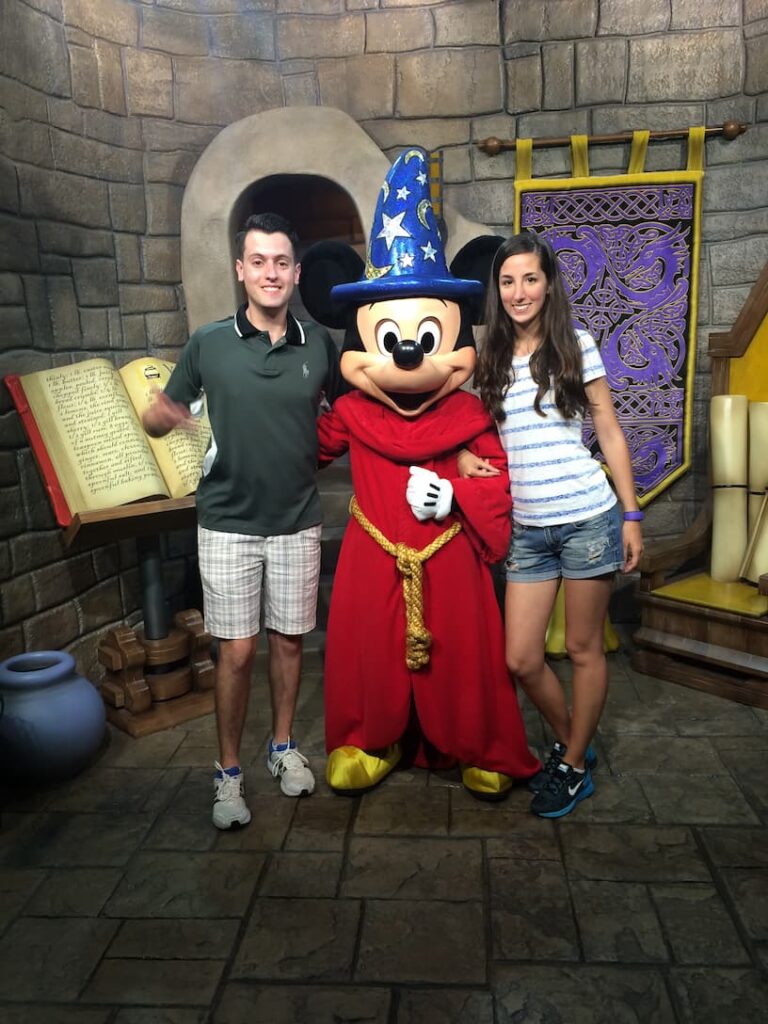 Jovem Casal posa junto ao Mickey Mouse vestido de feiticeiro, em um cenário que remete ao filme Fantasia, dentro do parque de diversões Hollywood Studios em Orlando, na Flórida