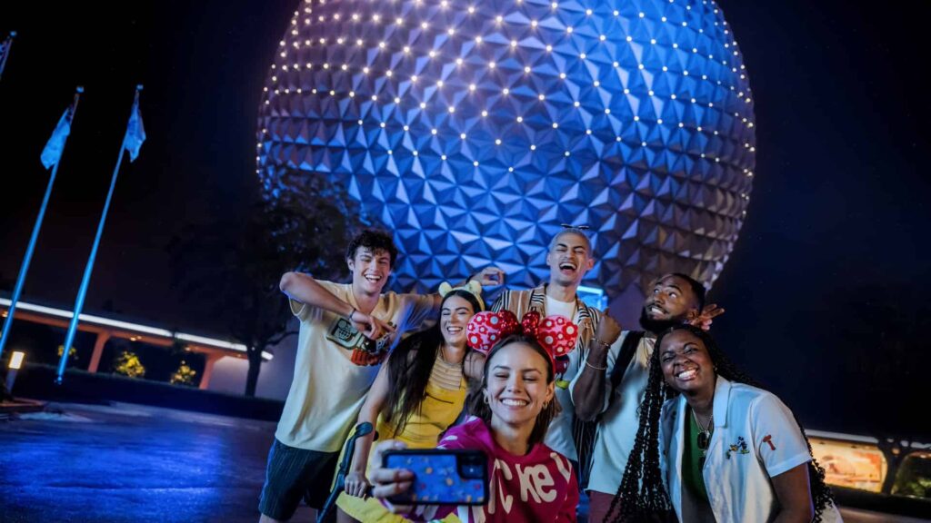 Grupo de pessoas felizes em frente a Spaceship Earth, a bola do Epcot, parque de diversões da Disney World, em Orlando na Flórida. Uma mulher a frente, com orelhinhas da Minnie, tem o celular posicionado para tirar uma selfie dela com os amigos durante o Evento After Hours, os amigos mais ao fundo parecem muito animados