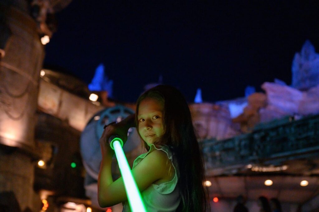 Garotinha aponta um sabre de luz verde em direção a tela, ao fundo temos Star Wars Land, Batuu, iluminada a noite, durante o Evento After Hours, da Disney World, no Hollywood Studios